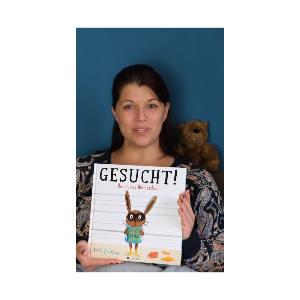 Frau hält ein Kinderbuch mit dem Titel „Gessucht! Henri, der Bücherdieb“ und neben ihr steht ein lächelnder Teddybär.